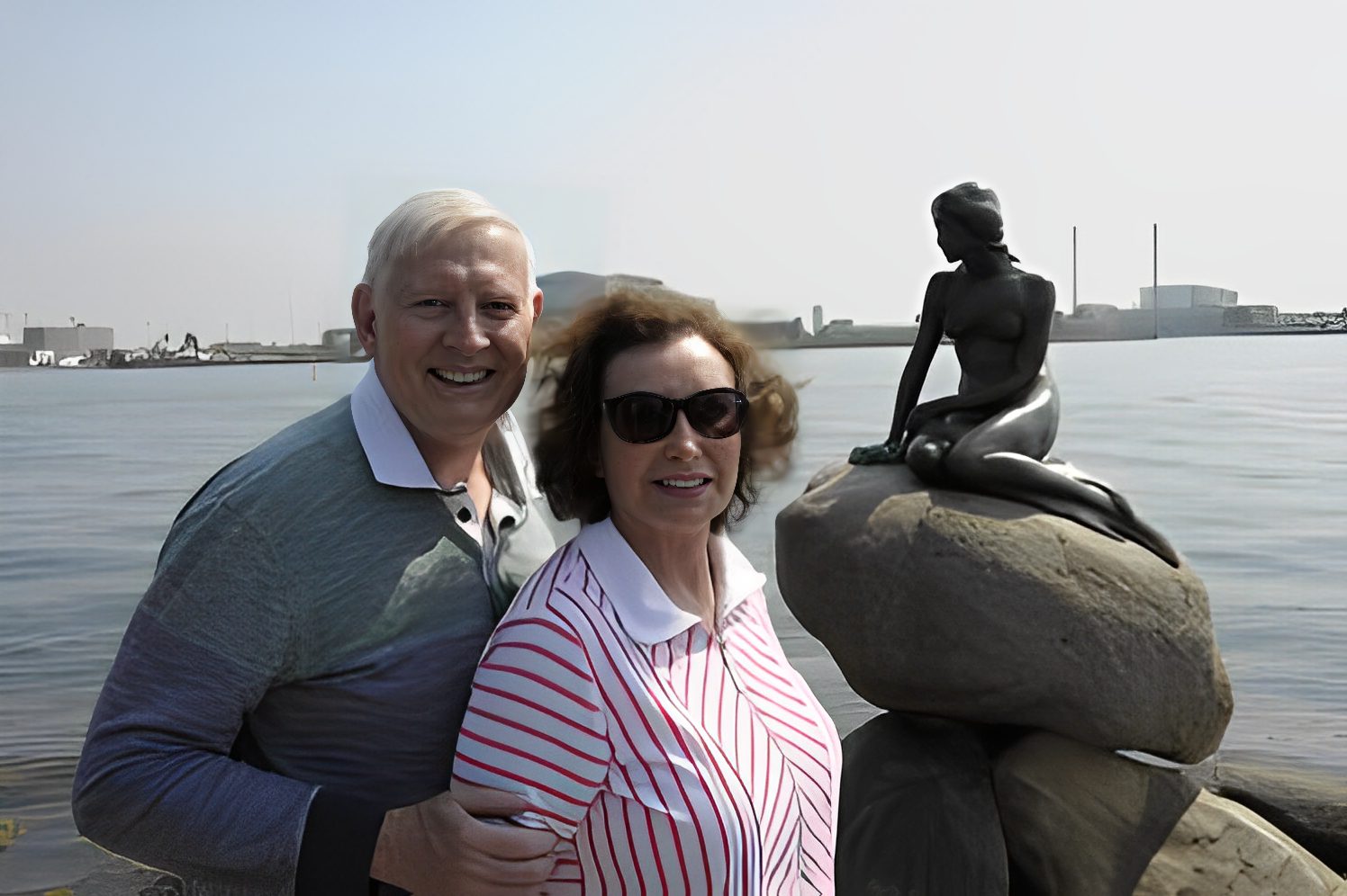 Couple smiling beside mermaid statue by water.