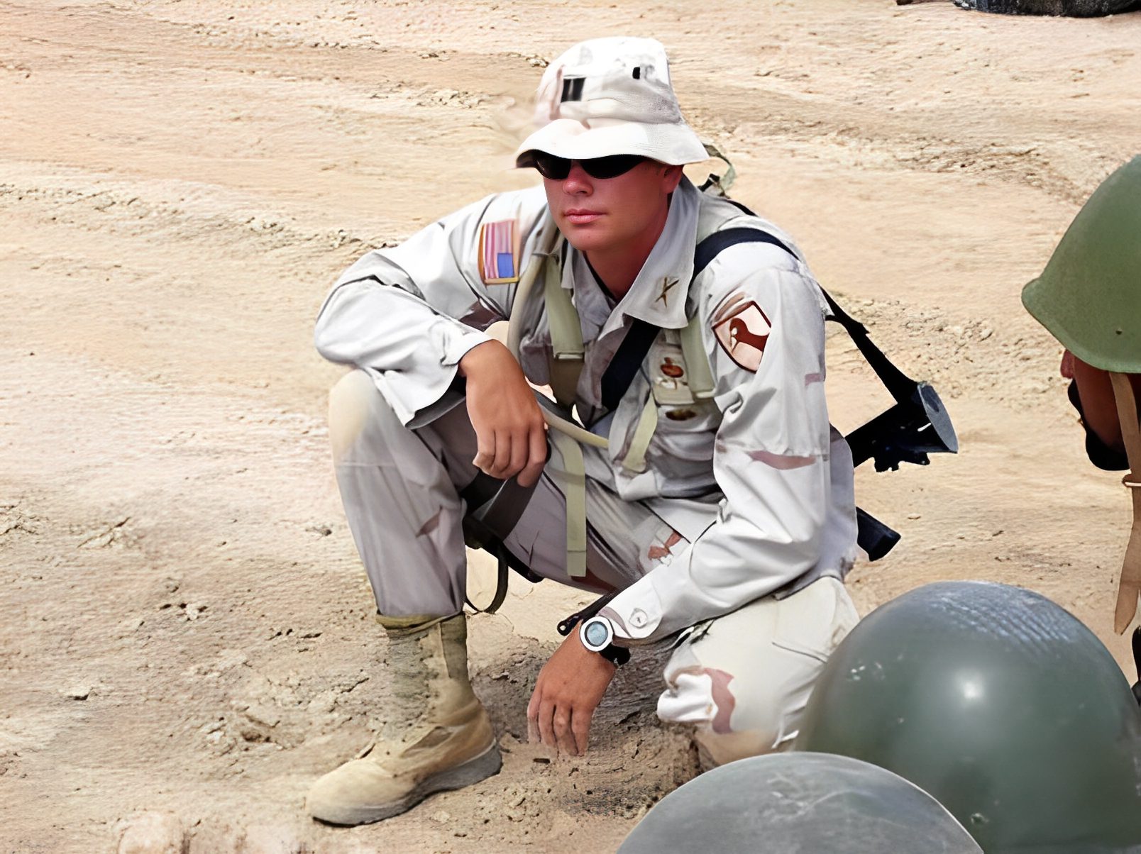 Soldier squatting on sandy ground, wearing uniform.